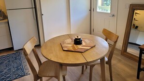 Una mesa redonda de madera con un hervidor negro, rodeada de sillas de madera, en una habitación con una puerta blanca y una ventana.