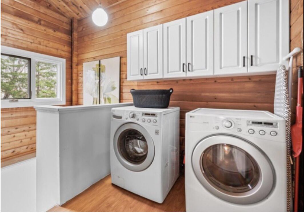 A laundry area with two washing machines, wooden cabinets, and a window with a view of trees.