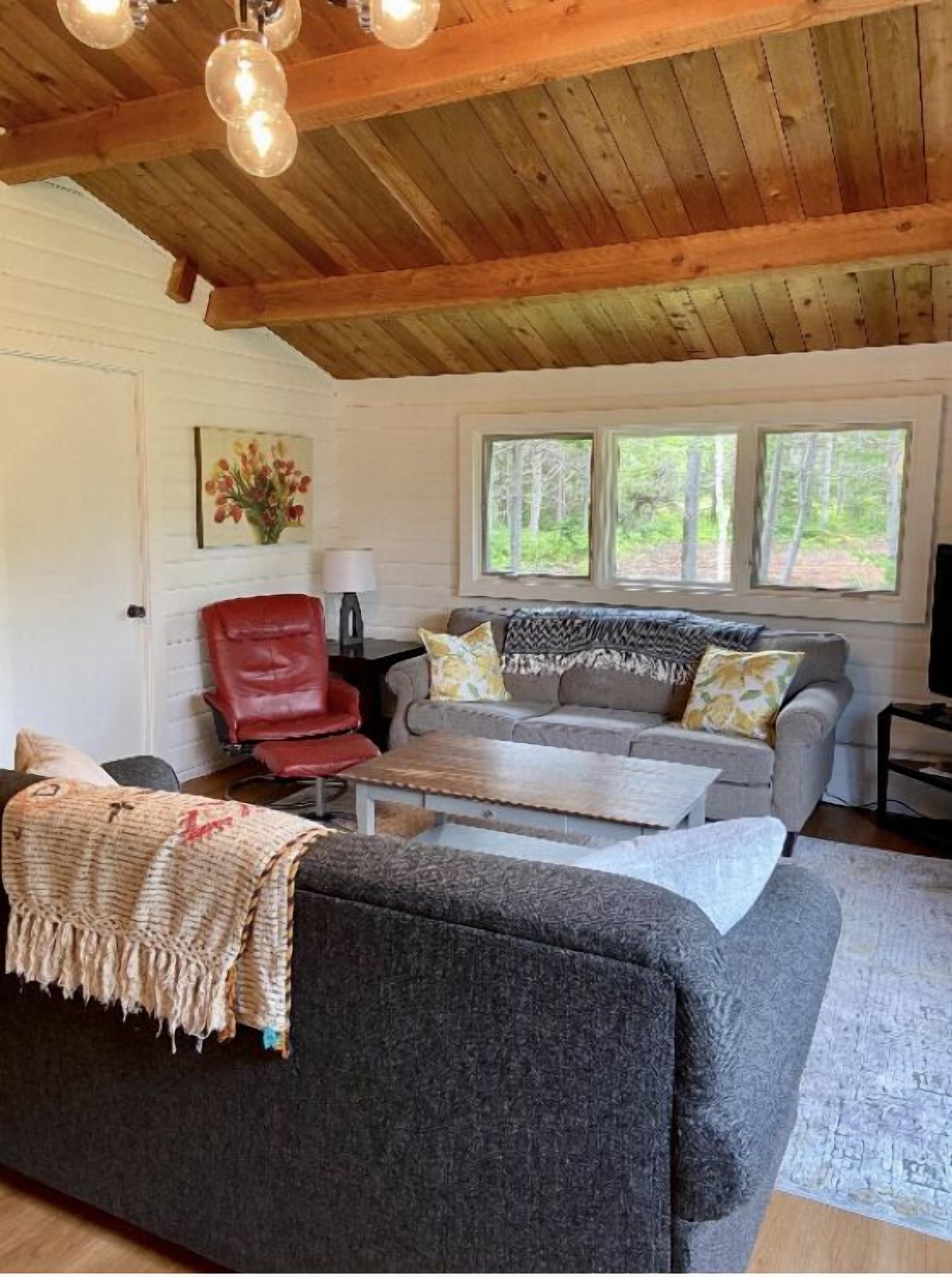 A cozy living room with a grey sofa, red chair, and a coffee table. There are two windows with a view of trees outside.
