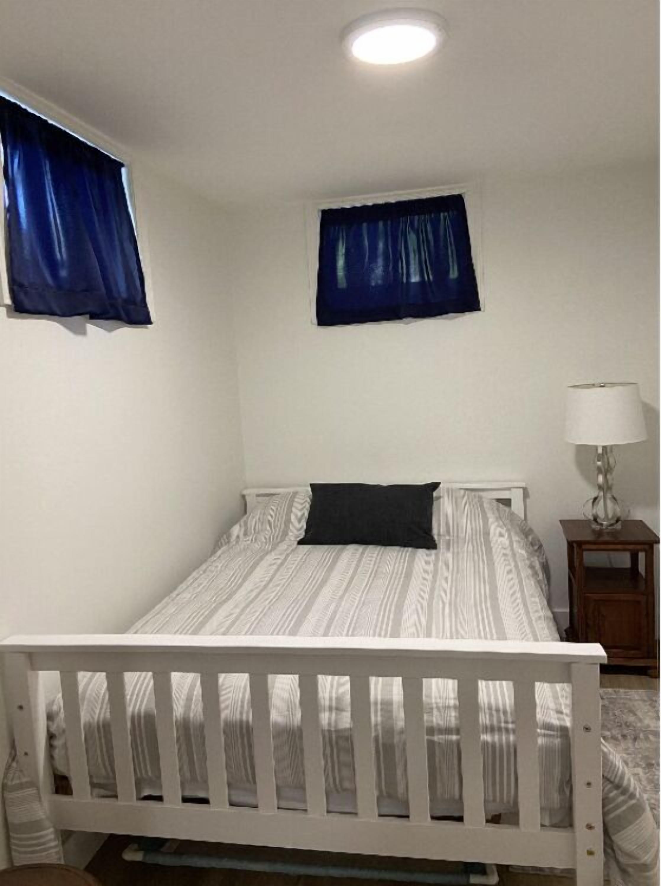 Una piccola camera con un letto singolo, un telaio bianco, un comodino con una lampada e due finestre con tende blu.