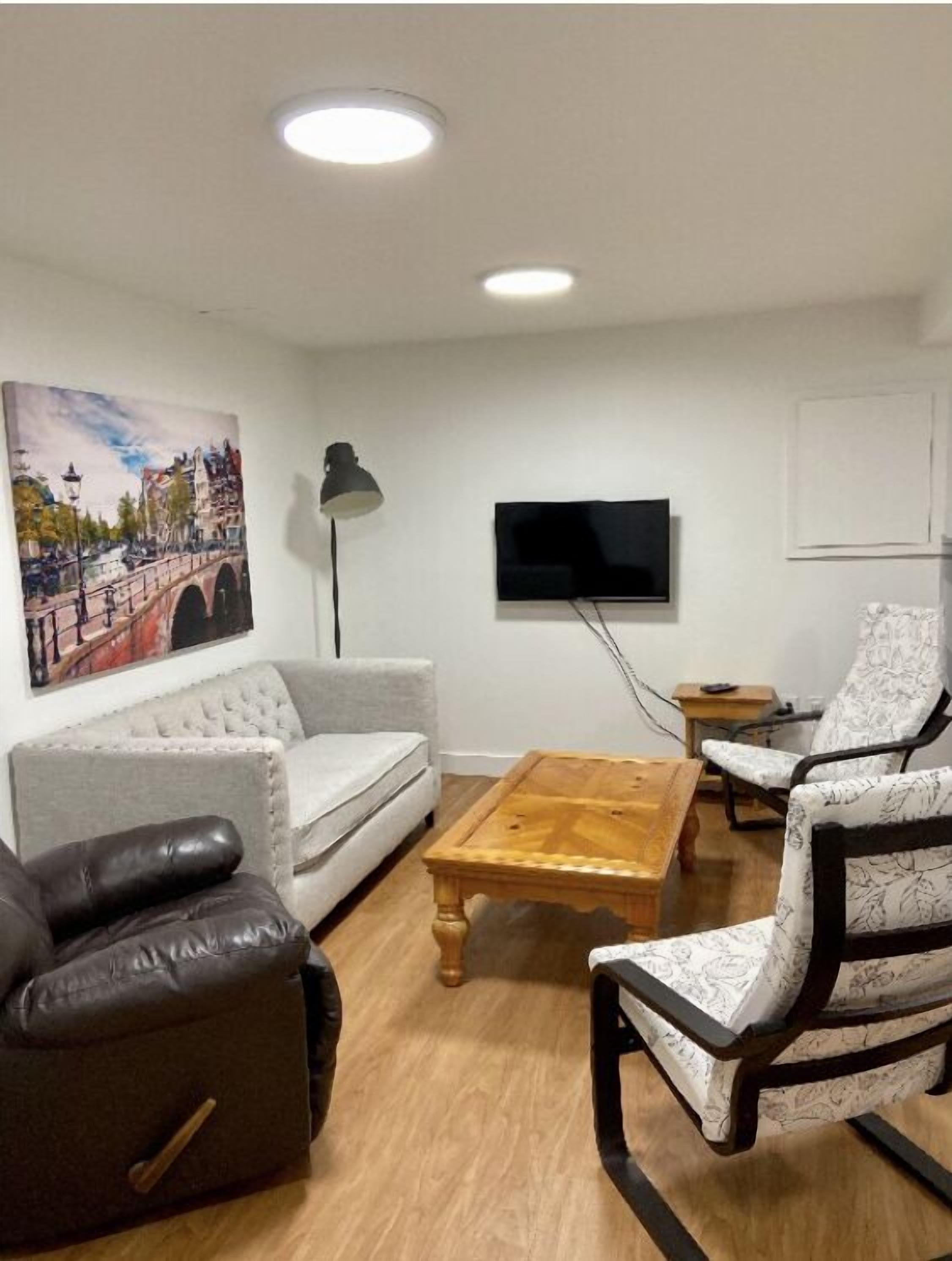 Ein Wohnzimmer mit einer Couch, einem Couchtisch und zwei Sesseln. An der Wand hängt ein Gemälde, und darüber ein Fernseher.