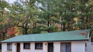 A small, single-story cabin with a green metal roof, white siding, and a porch with wooden benches. - Water Rafts + Trails - North Star by Janix Escapes. (Wellston)