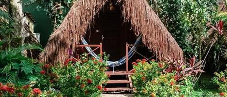 A thatched-roof hut with a hammock and a stone pathway leading to it, surrounded by lush greenery and flowering plants.