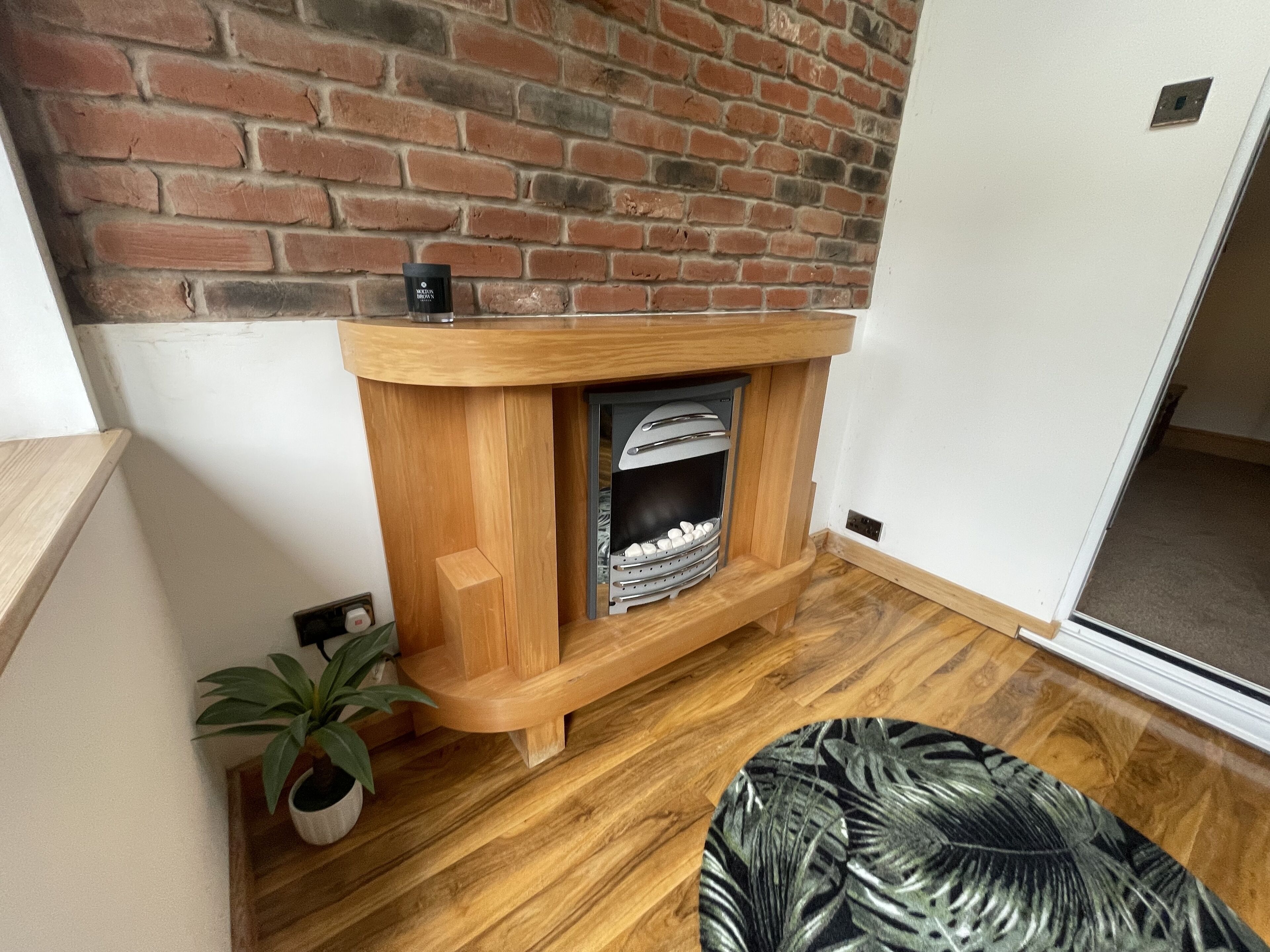 A corner room with a brick wall, a wooden fireplace, and a potted plant.
