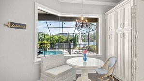 Una habitación con un amplio ventanal con vista a una piscina, una mesa de comedor blanca, un sillón estampado y una silla de mimbre.