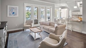 Una sala de estar amplia con un gran ventanal, un sofá, una mesa de centro y una zona de cocina con armarios blancos.