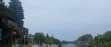 A wooden boardwalk with white railings, a river, and buildings on the opposite bank.