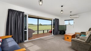 A spacious living room with a large sliding glass door leading to a balcony, a television, a sofa, and a wooden side table. - LAKE VIEW RETREAT - Relax, Unwind, & Make Memories (Mangakino)