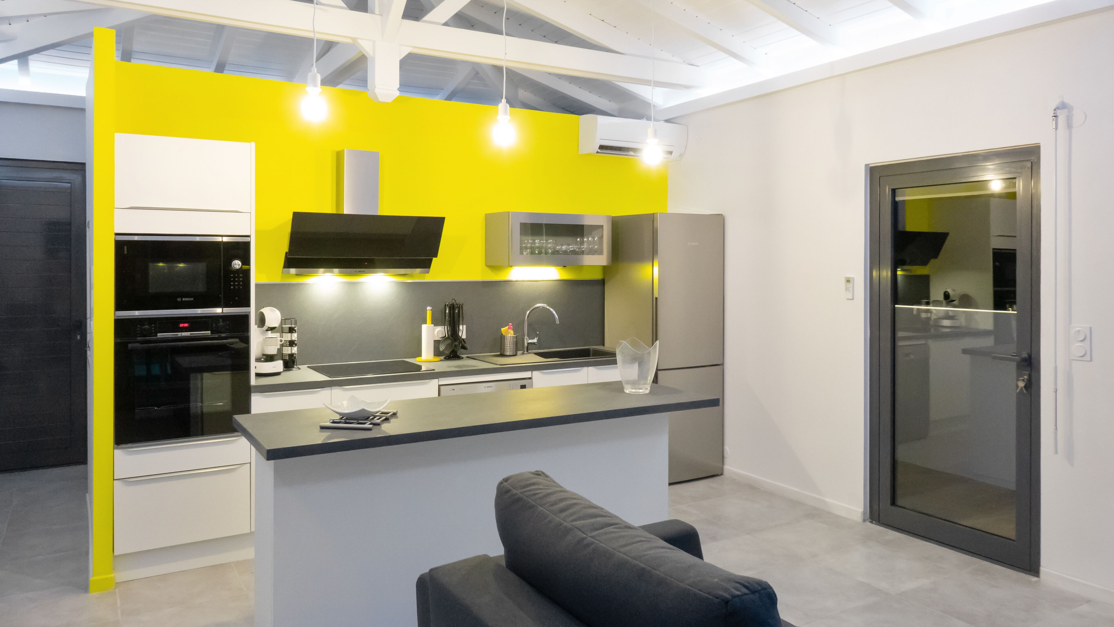Une cuisine moderne avec un mur d’accent jaune, des appareils électroménagers intégrés et un canapé gris.