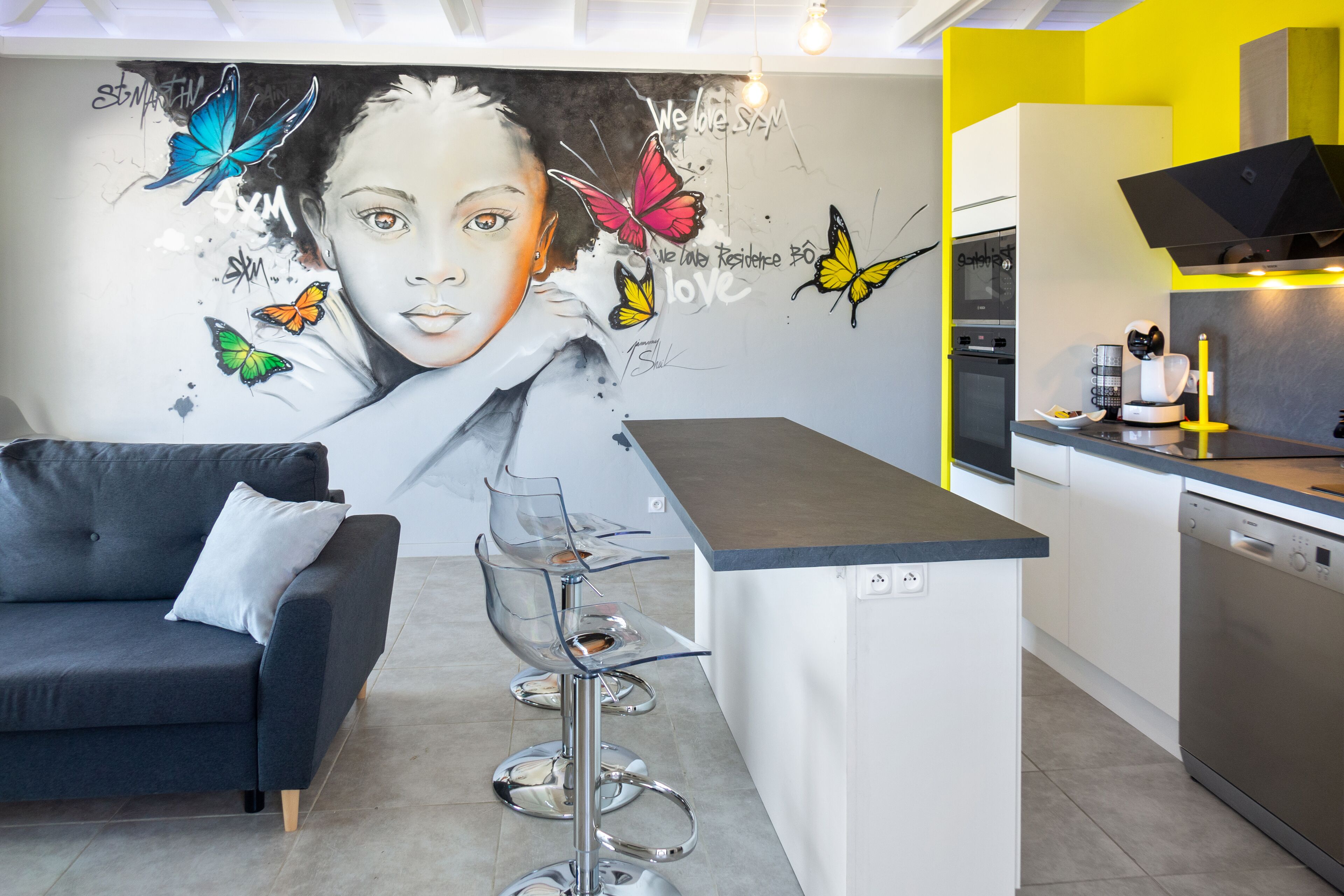 A modern kitchen with a grey sofa, a bar stool, and a mural with butterflies and text.