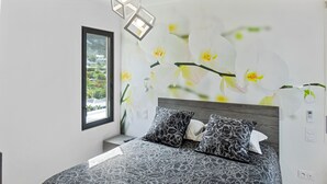 A modern bedroom with a large bed, two patterned pillows, a nightstand, a wall-mounted lamp, and a window with a view of greenery. - Residence BO, One-Bedroom Apartment, Suite with Sea View (Baie Orientale)