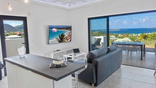 A modern kitchen with a dining area, a large window with a view of the ocean, and a flat-screen TV mounted on the wall. - Residence BO, One-Bedroom Apartment, Suite with Sea View (Baie Orientale)