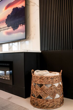 Een moderne woonkamer met een open haard, een aan de muur bevestigde televisie en een rieten poef met kussens.