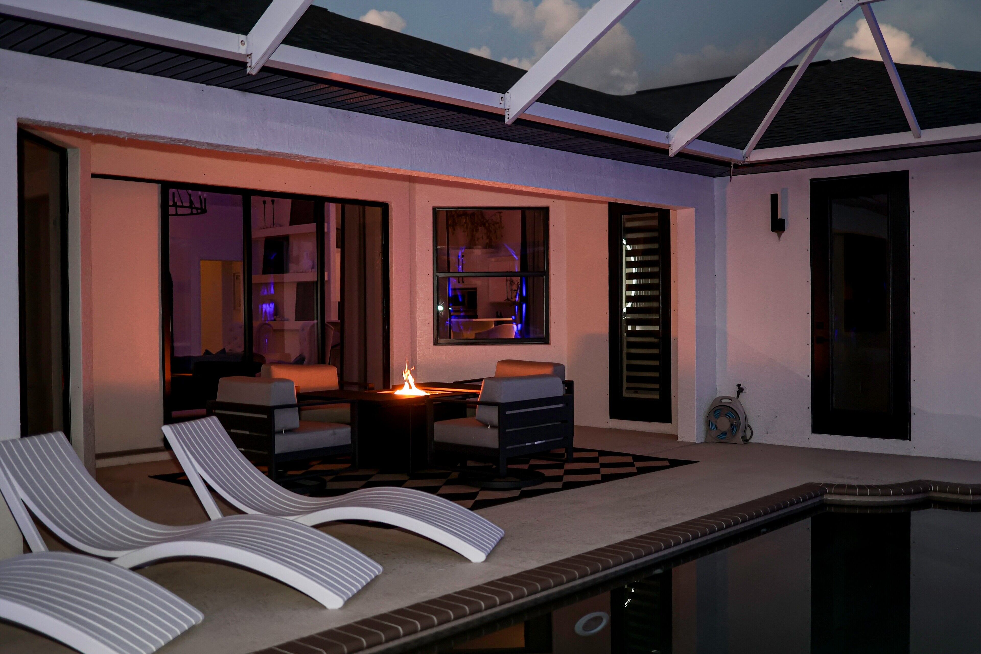 Un espace aménagé au bord de la piscine, avec des chaises longues, un foyer et une vue sur l’intérieur.