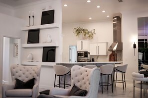 Een moderne woonkamer met een bank en stoelen, een keuken met witte kasten en apparaten, en een decoratieve plant.