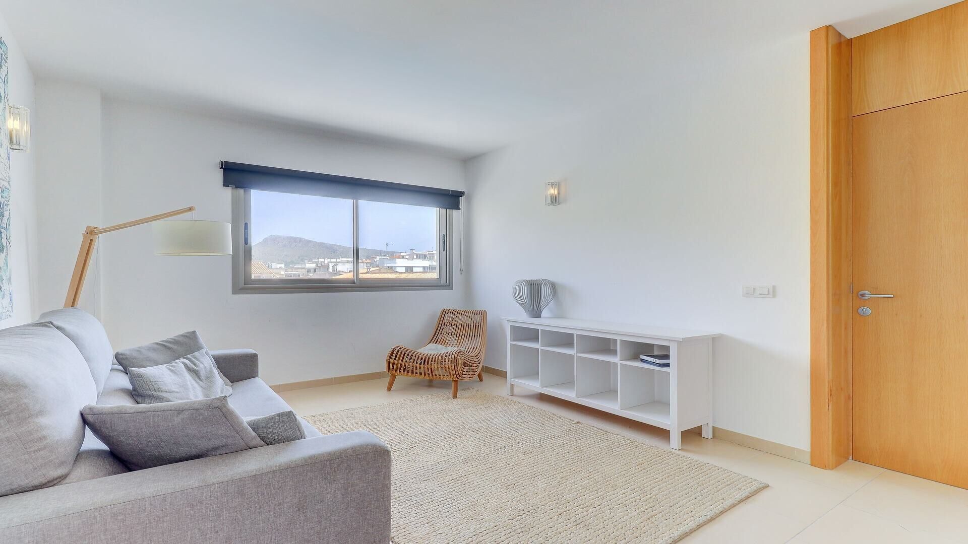 A modern living room with a grey sofa, a wicker chair, a white console table, and a large window with a view of buildings.