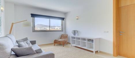 A modern living room with a grey sofa, a wicker chair, a white console table, and a large window with a view of buildings.