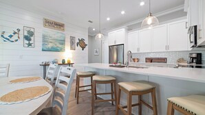 Une cuisine moderne avec des meubles blancs, un îlot central doté de tabourets et des œuvres d’art murales sur le thème de la marine.