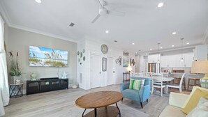 Un salon spacieux avec une table basse en bois, un fauteuil bleu et un canapé beige. On y trouve également un coin repas avec une table et des chaises blanches, ainsi qu’une cuisine dotée de meubles blancs et d’appareils électroménagers en acier inoxydable.