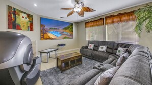 A living room with a gray sofa, a coffee table, a ceiling fan, a painting of a pool, and a large window with blinds.