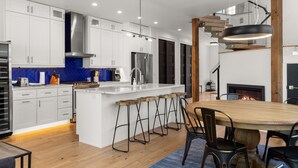 A modern kitchen with white cabinets, a central island with stools, a stainless steel refrigerator, and a wooden dining table with chairs. - Impressive, brand new modern residence in Whitefish. Close to downtown, lake and ski bus! (Whitefish)