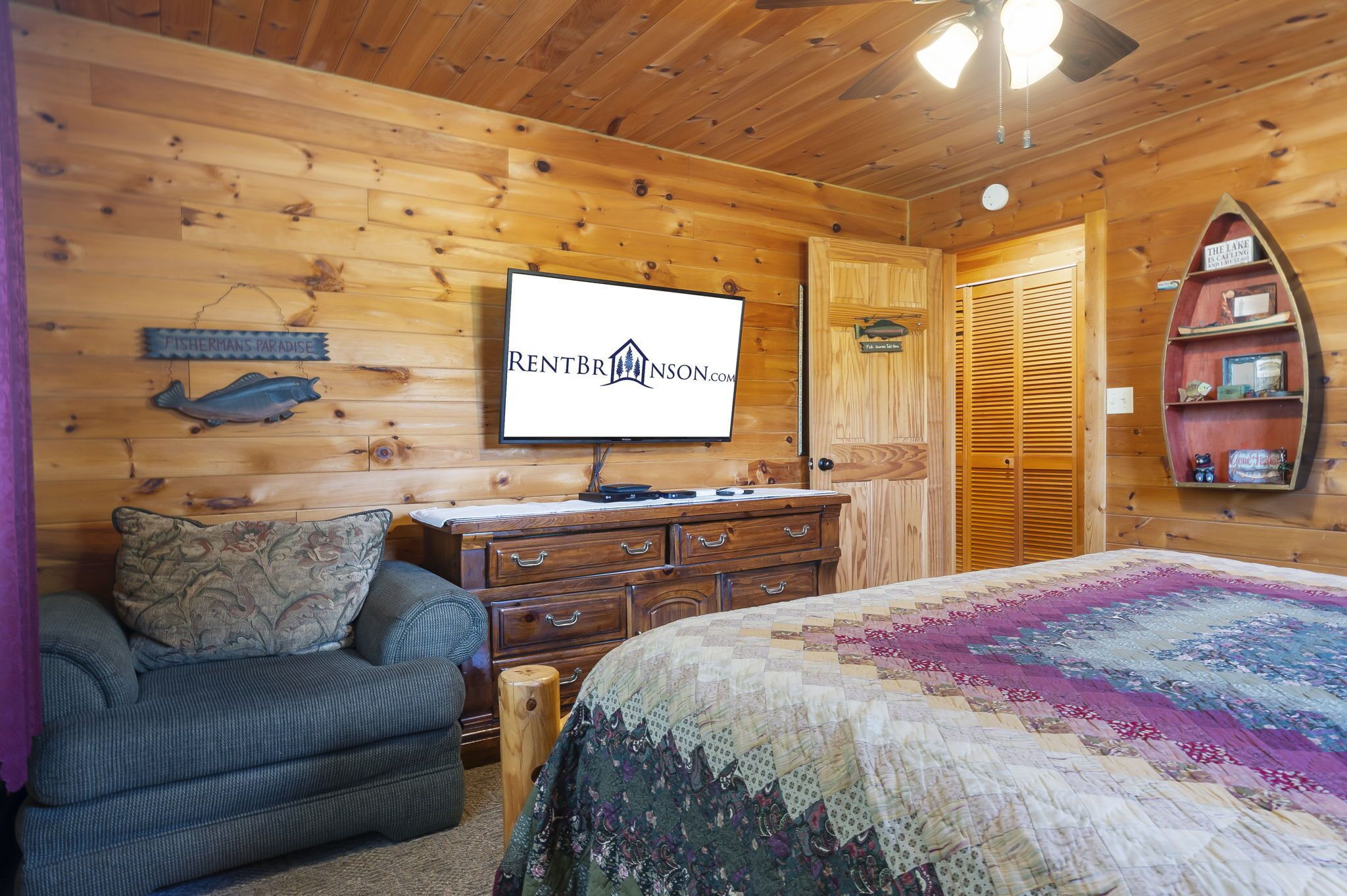 A cozy cabin room with a wooden interior, a bed with a colorful quilt, a dresser, a television, and a shelf with decorative items.