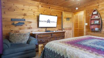 A cozy cabin room with a wooden interior, a bed with a colorful quilt, a dresser, a television, and a shelf with decorative items.