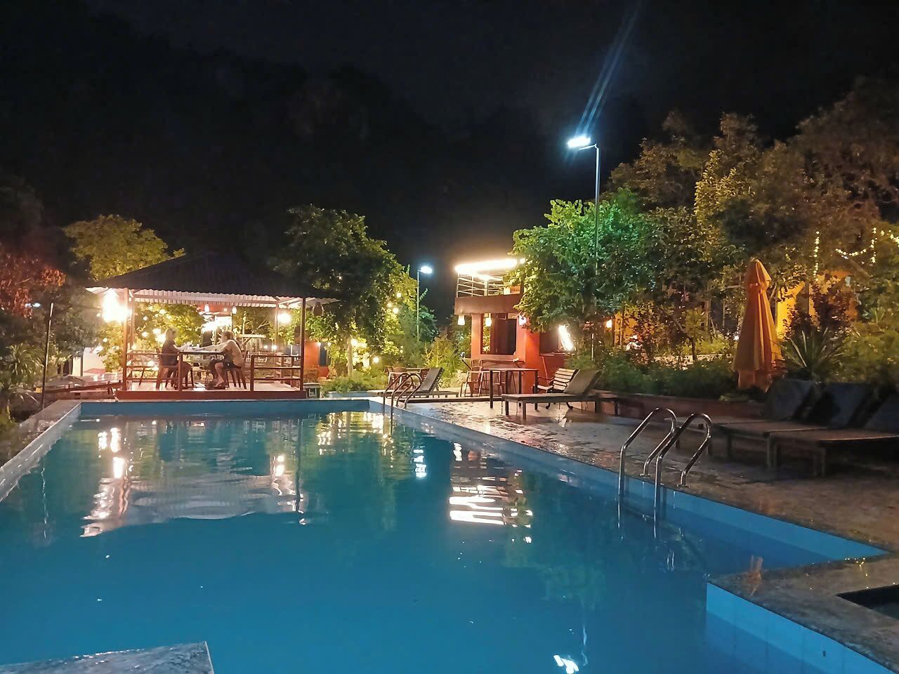 A swimming pool with lounge chairs and tables, surrounded by trees and a building with outdoor lighting.