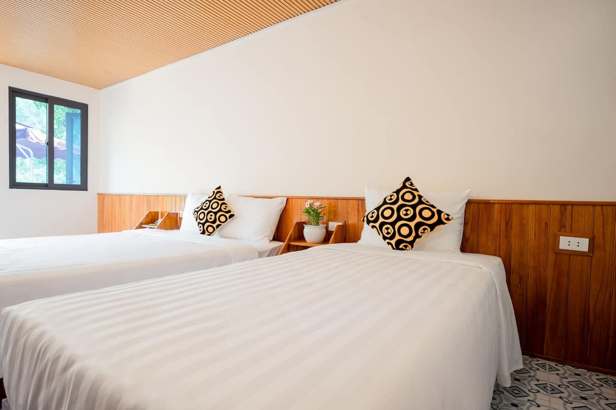 Habitación de hotel con dos camas, cabeceros de madera, ropa de cama blanca, almohadas estampadas, una pequeña planta en maceta y una ventana con vistas.