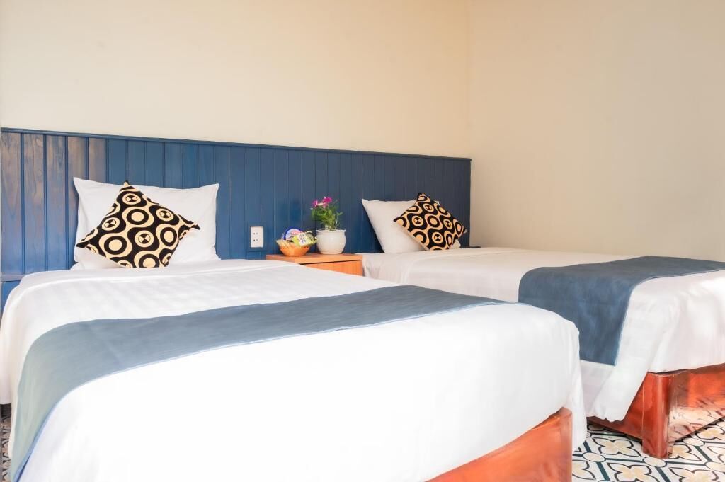 Ein Hotelzimmer mit zwei Betten, jeweils mit einer weißen und blauen Tagesdecke und gemusterten Kissen. Es gibt einen kleinen Tisch mit einer Pflanze und einem Obstkorb.
