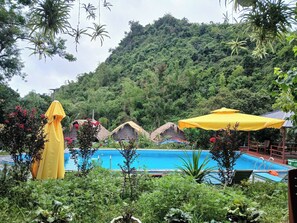 Un área de piscina con una vista despejada de una montaña al fondo, rodeada de exuberante vegetación y cabañas con techo de paja.