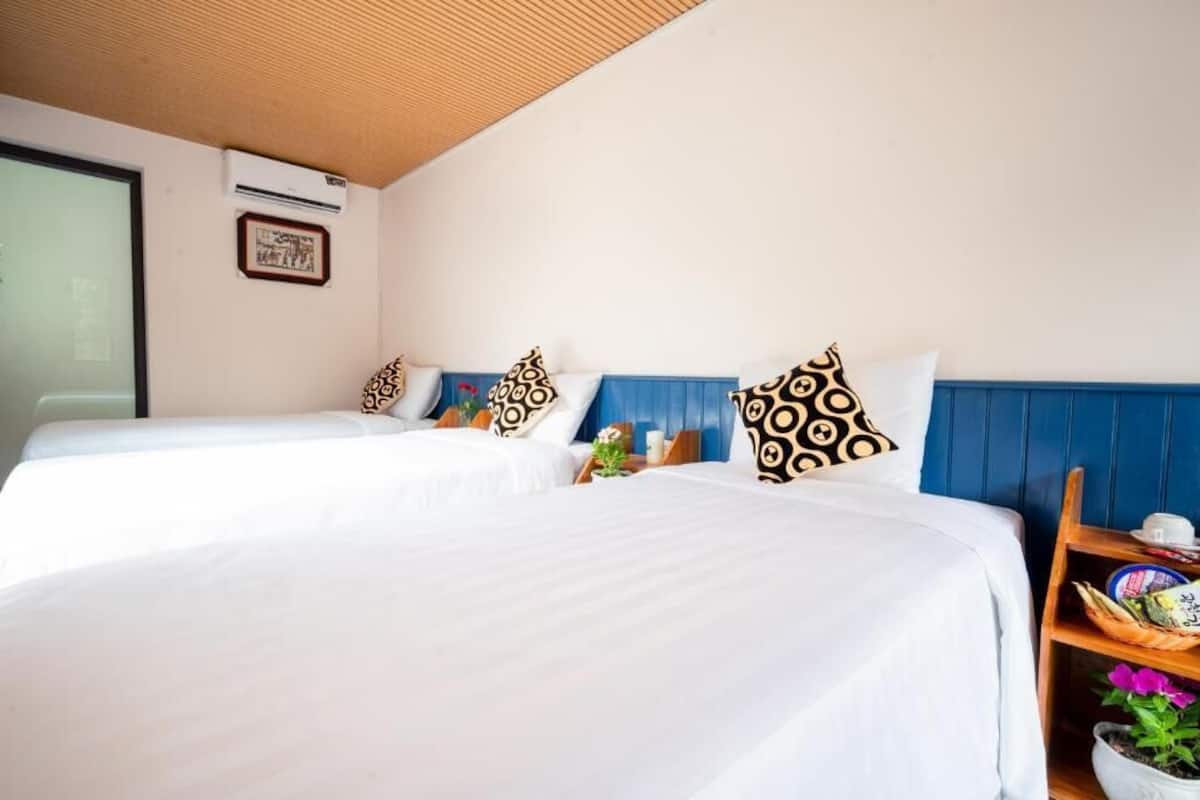 Habitación de hotel con dos camas, almohadones estampados, cabecera azul, mesita de madera con una taza y una cesta, y una planta en maceta.
