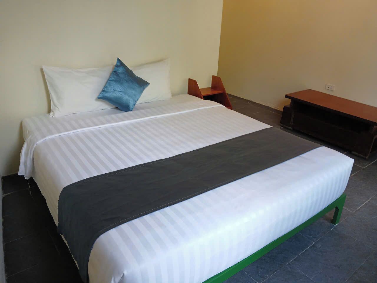 Een netjes opgemaakt bed met een grijs-wit gestreept dekbed, een blauw kussen en een donkergrijze plaid. Tegen de muur staat een houten bankje en een klein tafeltje met een donker oppervlak.