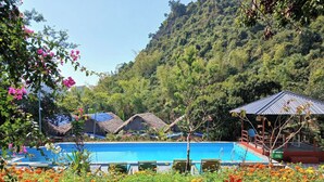 A pool area with lounge chairs, a thatched-roof structure, and a hill with dense vegetation in the background.