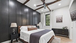 Une chambre avec un grand lit, des murs sombres, un ventilateur de plafond, une fenêtre et une commode.