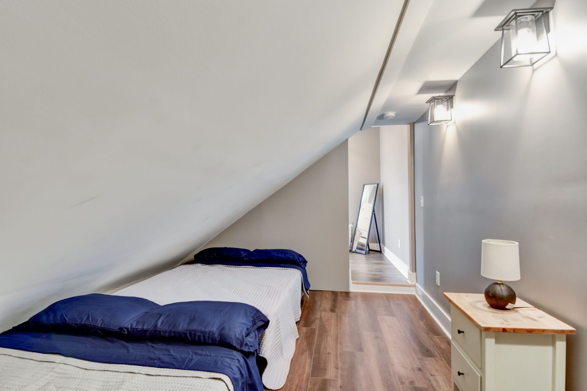 Una camera da letto con soffitto spiovente, un letto con biancheria azzurra, un comodino con una lampada e un corridoio con uno specchio e un quadro incorniciato.