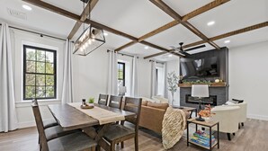 A modern living room with a dining area, wooden beams on the ceiling, a fireplace, and a TV mounted on the wall.