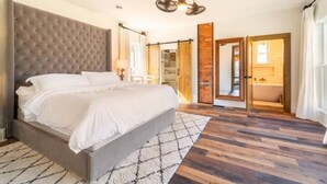 Un dormitorio con una cama grande, piso de madera, ventilador de techo y baño visible a través de una puerta abierta.
