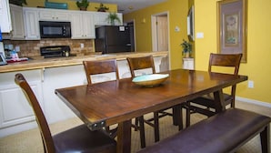 A dining area with a wooden table and chairs, a kitchen in the background, and a yellow wall.