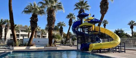 A swimming pool with a water slide, palm trees, and a clear blue sky.
