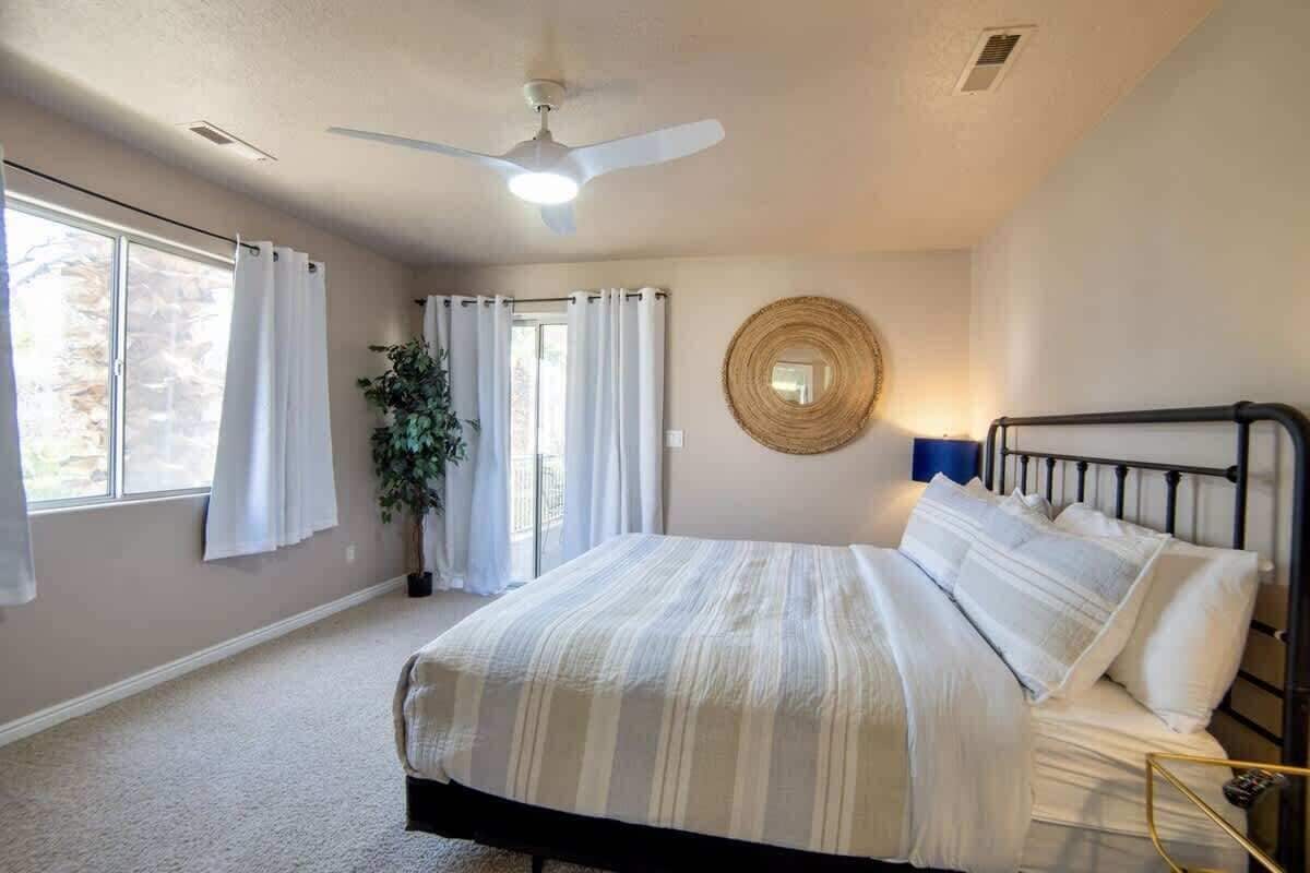 A bedroom with a bed, a ceiling fan, a window with curtains, and a plant.