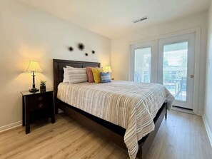 A bedroom with a bed, two bedside tables, a window with blinds, and a view of the outdoors.