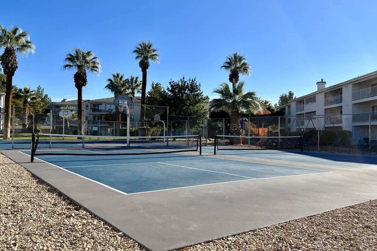 Una cancha de tenis al aire libre con red, rodeada por una cerca y palmeras.