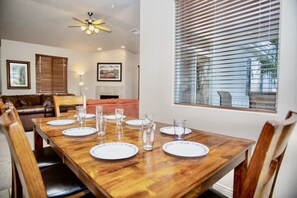 A dining area with a wooden table set for four, surrounded by chairs, with a ceiling fan above and a window with blinds to the right.