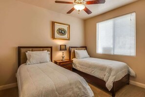 A bedroom with two beds, a ceiling fan, a bedside lamp, a framed picture on the wall, and a window with blinds.