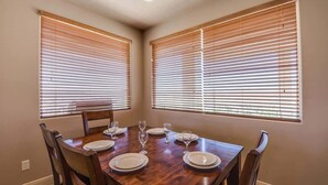 A dining room with a wooden table set for four, flanked by two windows with blinds.