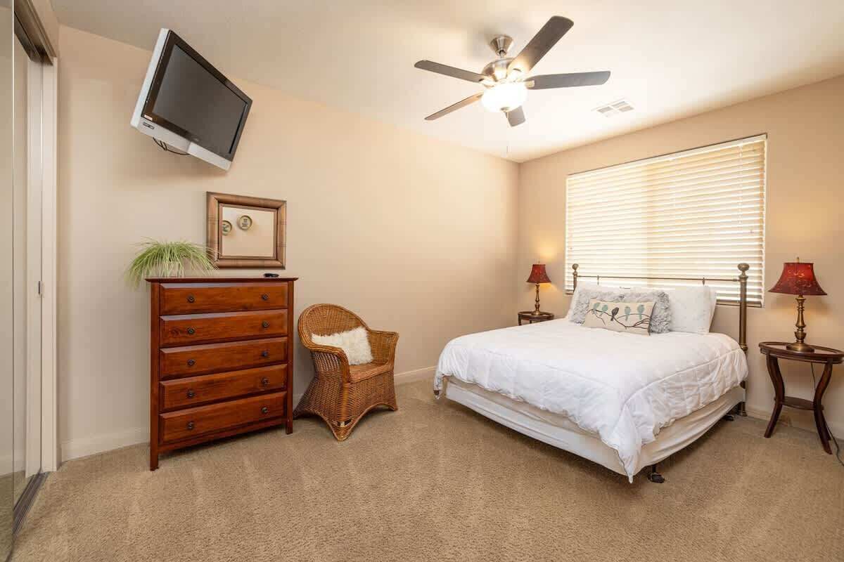 Une chambre à coucher comprenant un lit, une commode, une chaise, un ventilateur de plafond, un téléviseur et des lampes de chevet.