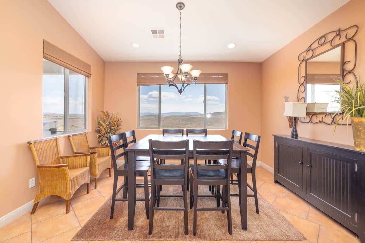 Un coin repas comprenant une table et des chaises, un lustre, une grande fenêtre donnant sur un beau paysage et un meuble avec un miroir.