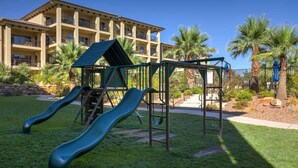 A playground with two slides and a climbing structure, surrounded by grass and palm trees, with a building in the background.