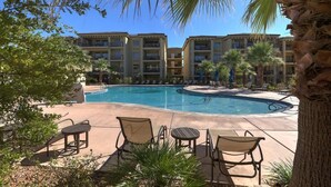 A swimming pool with lounge chairs and tables, surrounded by palm trees and a building with balconies.